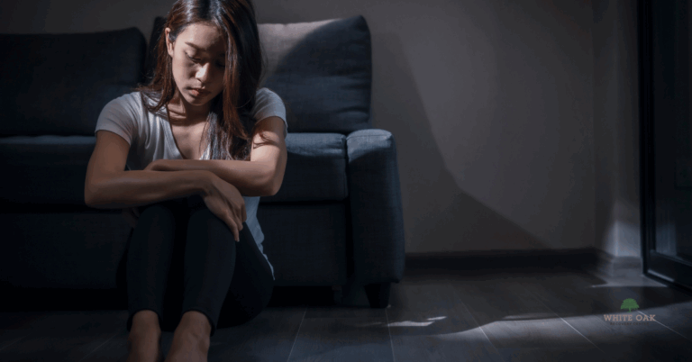 Young woman sitting on the floor in a dark room looking distressed, representing depression and co-occurring addiction treatment at White Oak Recovery Center